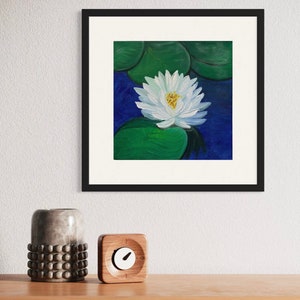 May include: A framed painting of a white water lily with a yellow center, surrounded by green lily pads, set against a deep blue background. The artwork is displayed in a black frame with a white mat. The painting is displayed on a wooden surface.