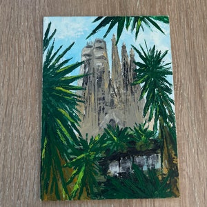 May include: An acrylic painting of the Sagrada Familia in Barcelona, Spain, framed by green palm fronds. The cathedral is depicted in shades of gray and beige against a light blue sky. The artwork is on a rectangular canvas.