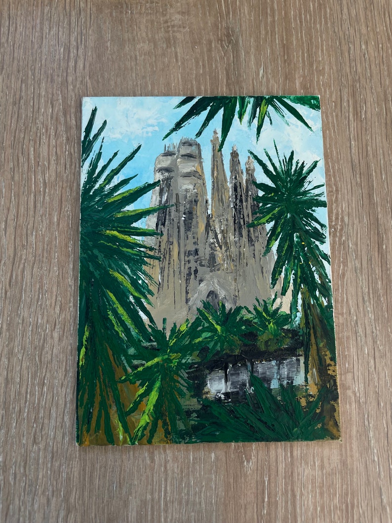 May include: An acrylic painting of the Sagrada Familia in Barcelona, Spain, framed by green palm fronds. The cathedral is depicted in shades of gray and beige against a light blue sky. The artwork is on a rectangular canvas.