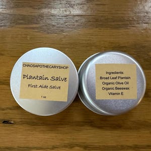 May include: Two round silver metal tins containing plantain salve. The left tin features a tan label with the text CHAOSAPOTHECARYSHOP, Plantain Salve, First Aide Salve, and 1 oz. The right tin has a tan label listing ingredients: Broad Leaf Plantain, Organic Olive Oil, Organic Beeswax, and Vitamin E.
