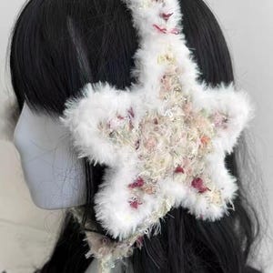 Puede incluir: Orejeras blancas y esponjosas con un diseño grande en forma de estrella. La estrella está adornada con una mezcla de acentos florales rosas y crema. La diadema y las orejeras están hechas de un material suave y afelpado.
