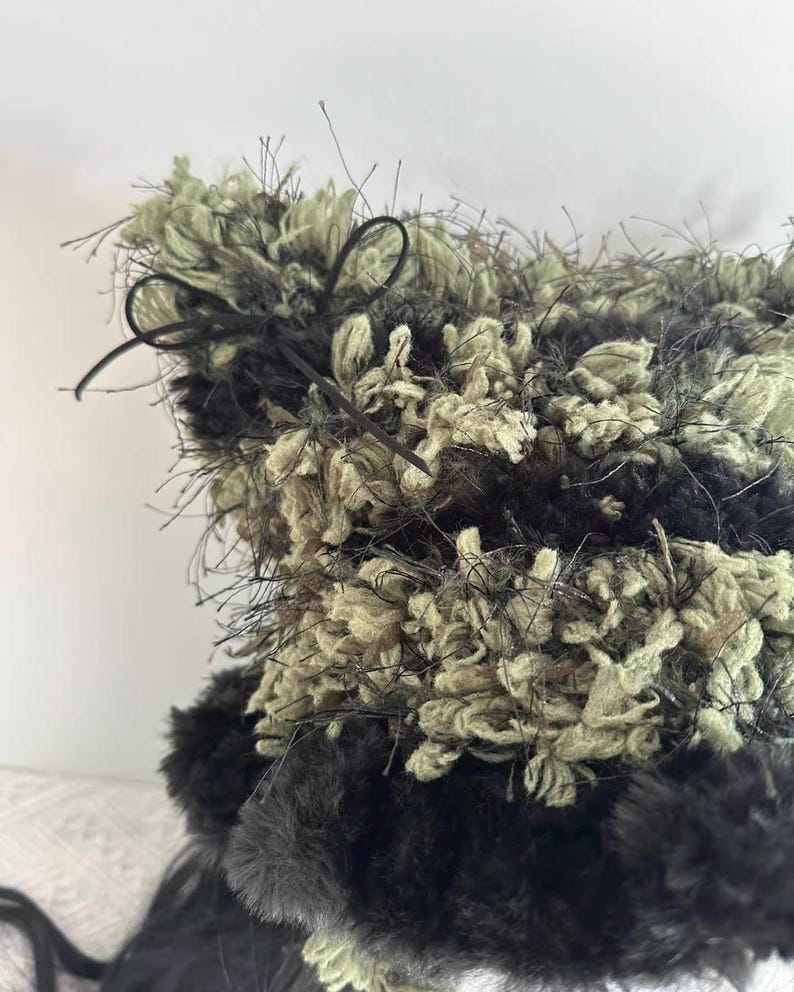 Puede incluir: Un sombrero texturizado hecho a mano con una mezcla de hilo negro, verde oliva y beige. El sombrero presenta un dise&ntilde;o esponjoso y peludo con un lazo de cinta negra. La textura y la paleta de colores &uacute;nicos del sombrero crean un accesorio elegante.