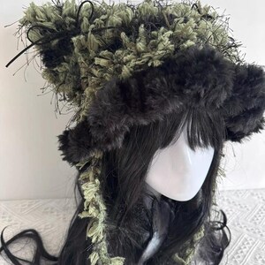 Puede incluir: Un tocado &uacute;nico con una combinaci&oacute;n de texturas y colores. El sombrero tiene un borde esponjoso de color gris oscuro y una secci&oacute;n superior con una mezcla de material difuso verde oliva y negro. Hebras y cintas negras a&ntilde;aden detalles.