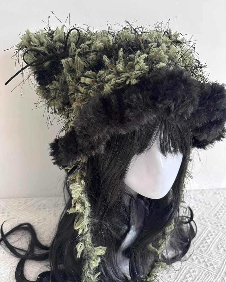 Puede incluir: Un tocado &uacute;nico con una combinaci&oacute;n de texturas y colores. El sombrero tiene un borde esponjoso de color gris oscuro y una secci&oacute;n superior con una mezcla de material difuso verde oliva y negro. Hebras y cintas negras a&ntilde;aden detalles.