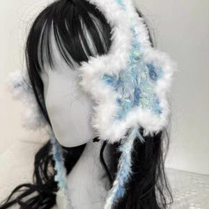Op de afbeelding: Witte en blauwe oorwarmers in de vorm van een ster met een zachte textuur. De oorwarmers hebben een hoofdband en stervormige oorbeschermers met blauwe en groene accenten. Lange, bungelende strengen van hetzelfde materiaal verbinden de oorbeschermers met de hoofdband.