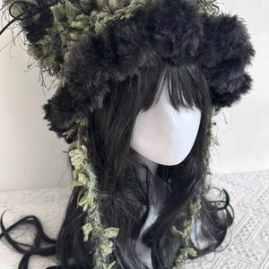 Puede incluir: Un tocado &uacute;nico con una combinaci&oacute;n de texturas y colores. El dise&ntilde;o incluye una banda esponjosa de color gris oscuro y una corona de material texturizado verde y negro. El pelo largo y oscuro enmarca la cara, con detalles verdes.