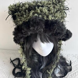 Puede incluir: Un sombrero &uacute;nico, hecho a mano, con una combinaci&oacute;n de texturas difusas verdes y negras. El sombrero tiene un borde oscuro y esponjoso alrededor de la cara y las orejas, con detalles negros y una corona texturizada. Accesorio de moda.