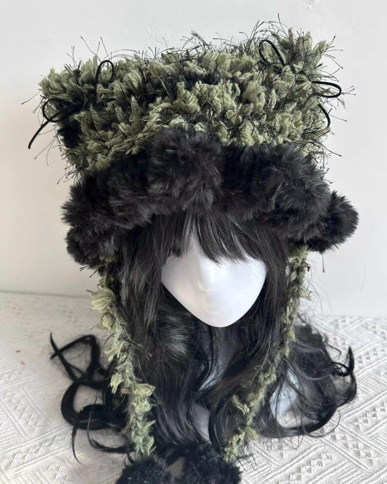 Puede incluir: Un sombrero &uacute;nico, hecho a mano, con una combinaci&oacute;n de texturas difusas verdes y negras. El sombrero tiene un borde oscuro y esponjoso alrededor de la cara y las orejas, con detalles negros y una corona texturizada. Accesorio de moda.