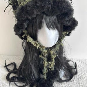Puede incluir: Un gorro de piel sint&eacute;tica negro y verde oliva con una bufanda y pompones a juego. El gorro tiene una textura esponjosa y est&aacute; dise&ntilde;ado para ser usado en la cabeza. La bufanda est&aacute; hecha de un material similar y est&aacute; unida al gorro.