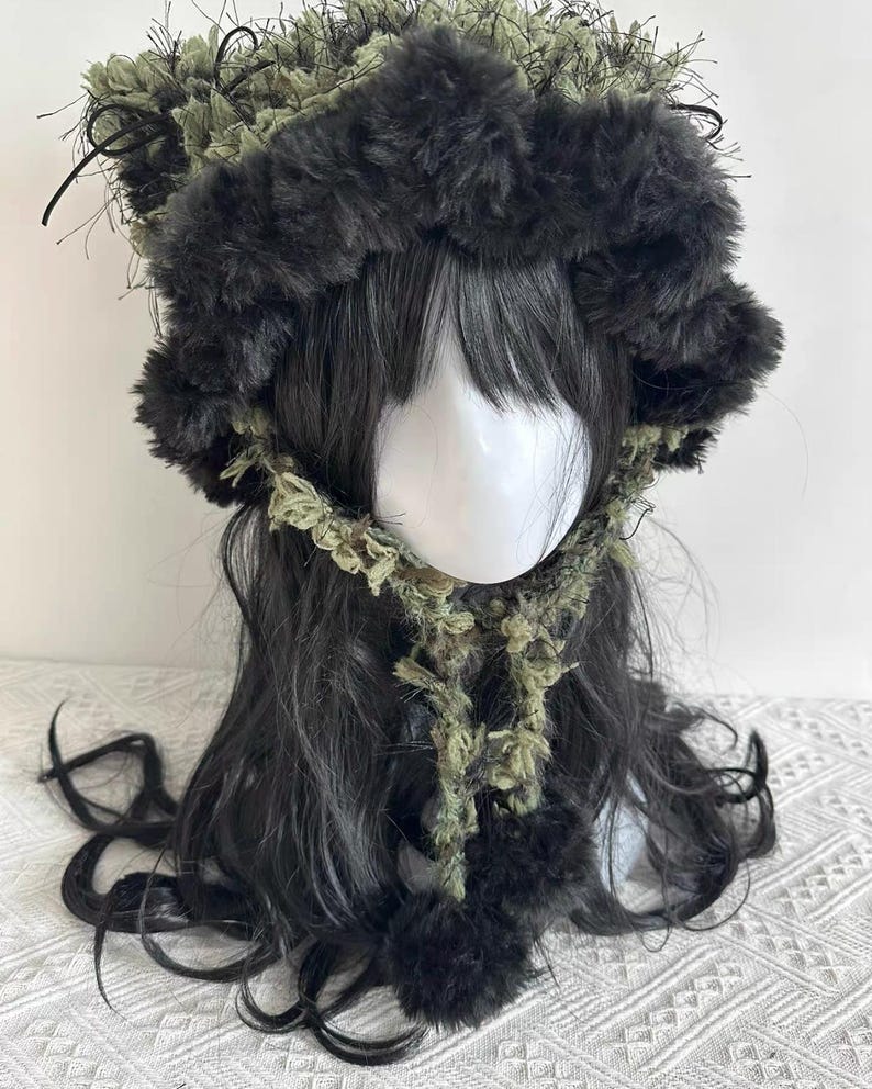 Puede incluir: Un gorro de piel sint&eacute;tica negro y verde oliva con una bufanda y pompones a juego. El gorro tiene una textura esponjosa y est&aacute; dise&ntilde;ado para ser usado en la cabeza. La bufanda est&aacute; hecha de un material similar y est&aacute; unida al gorro.