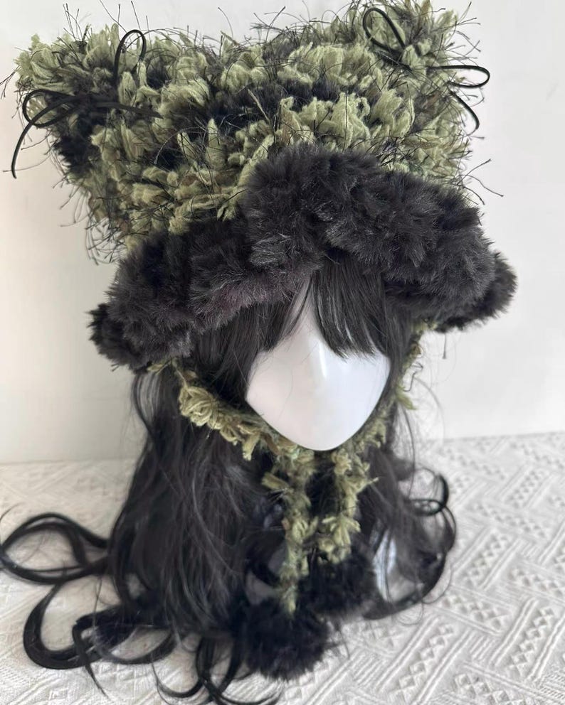 Puede incluir: Un sombrero caprichoso, hecho a mano, con un dise&ntilde;o &uacute;nico. El sombrero presenta una corona esponjosa y texturizada en tonos verdes y negros, con detalles en negro y peque&ntilde;os lazos. El ala es de un material m&aacute;s oscuro y afelpado. El sombrero forma parte de un conjunto de disfraz o cosplay.