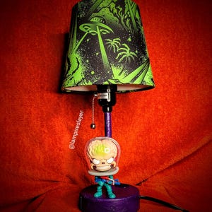 May include: A table lamp with a green and black lampshade featuring a UFO and palm trees. The lamp base is purple and features a pop culture figure of a creature with a brain in a helmet.