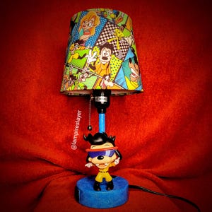 May include: A table lamp featuring a cartoon character with a blue and yellow outfit and a black and red mask. The lampshade is colorful and features a comic book style design with various cartoon characters. The lamp is on a red surface.