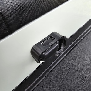 May include: A black plastic latch with a white label that says "12A MAX" is attached to a white surface. The latch is attached to a black surface with a textured pattern.