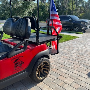May include: Red golf cart with a black roof and seats, a black metal bed, and a black and gold wheel. The cart has a sticker on the side that says "Skurrrits Customs" with a phone number and Instagram handle. An American flag is attached to the back of the cart.