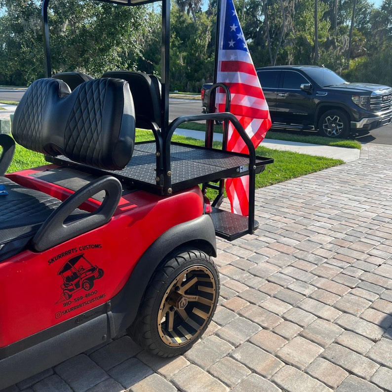 May include: Red golf cart with a black roof and seats, a black metal bed, and a black and gold wheel. The cart has a sticker on the side that says "Skurrrits Customs" with a phone number and Instagram handle. An American flag is attached to the back of the cart.