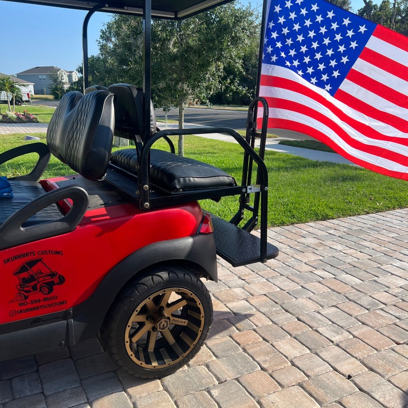May include: Red golf cart with black seats and a black roll bar. The cart has a black step and a black and gold wheel. The cart has a sticker on the side that says "Skurrats Customs" and a phone number. An American flag is in the background.