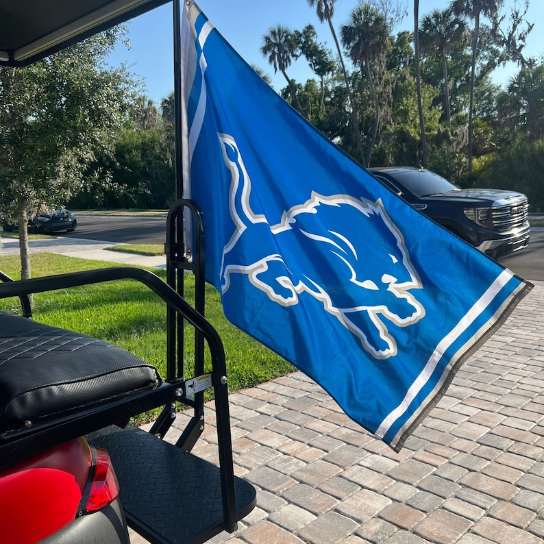 May include: A black golf cart with a blue and white Detroit Lions flag attached to the back. The flag has a white stripe at the bottom and a white stripe near the top. The flag features a blue lion with a white outline.