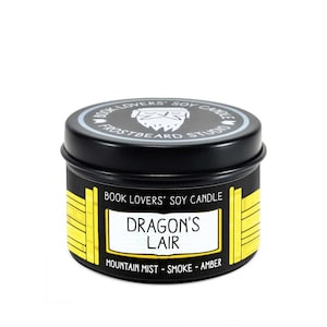 May include: A black soy candle tin with a lid featuring the text "BOOK LOVERS' SOY CANDLE" and "FROSTBEARD STUDIO". The label reads "DRAGON'S LAIR" with "MOUNTAIN MIST - SMOKE - AMBER" below. The tin has yellow accents.