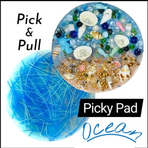 Può includere: Due opere d'arte rotonde in resina. Una presenta un design blu a forma di ragnatela. L'altra ha un tema spiaggia con conchiglie, perle e perline. Il testo sull'immagine recita "Pick & Pull", "Picky Pad" e "Ocean".