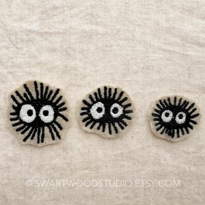 May include: Three embroidered patches of black and white fuzzy creatures with large eyes. The patches are on a beige fabric background.
