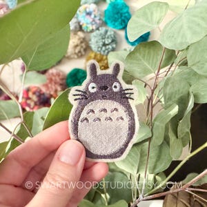 May include: A gray embroidered iron-on patch featuring a cartoon character with large ears and a fluffy body. The character has a friendly expression and is known as Totoro from the animated film My Neighbor Totoro.