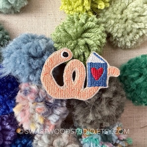 May include: An embroidered patch featuring a peach-colored worm shape with a black eye, holding a blue book with a red heart on the cover. The patch is surrounded by various colorful yarn pom-poms in shades of blue, green, and purple.