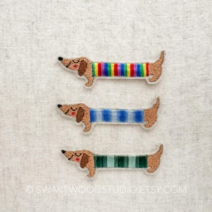 May include: Three embroidered dachshund patches with different colored striped backs. One has a rainbow stripe, another has blue stripes, and the third has green stripes. The dachshunds are brown with closed eyes, and white outlines.