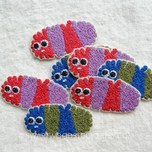 May include: A collection of embroidered patches shaped like segmented caterpillars with cartoon faces. The patches feature vibrant color combinations including red, purple, blue, and green, with black and white eyes and small stitched mouths.