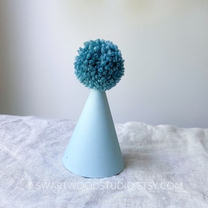 May include: A light blue party hat with a blue pom-pom on top. The hat is sitting on a white fabric surface.