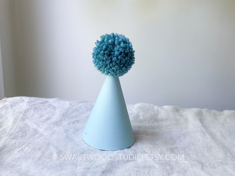 May include: A light blue party hat with a blue pom-pom on top. The hat is sitting on a white fabric surface.