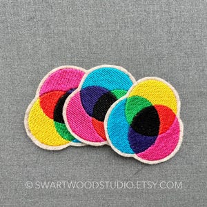 May include: Three colourful embroidered patches in the shape of overlapping circles. The patches are yellow, red, green, blue, pink and black.