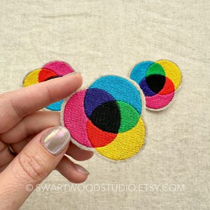 May include: Three colourful embroidered patches in the shape of circles, each with a different colour combination of overlapping circles. The colours are red, yellow, blue, green, purple, and black.