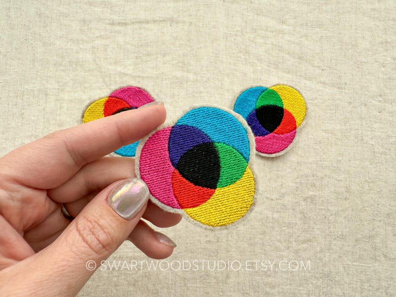 May include: Three colourful embroidered patches in the shape of circles, each with a different colour combination of overlapping circles. The colours are red, yellow, blue, green, purple, and black.