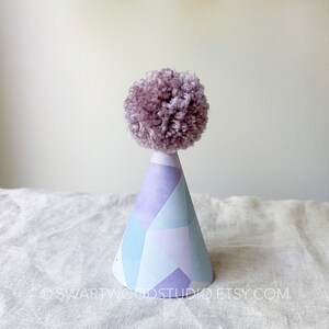 May include: A purple pom-pom sits atop a blue and purple geometric patterned party hat.