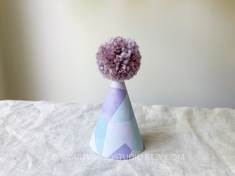 May include: A purple pom-pom sits atop a blue and purple geometric patterned party hat.