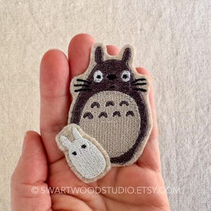 Może przedstawiać: Naszywka haftowana w kolorze brązowym i szarym przedstawiająca Totoro, postać z filmu animowanego M&oacute;j sąsiad Totoro, z mniejszą, białą haftowaną naszywką przedstawiającą kr&oacute;lika.