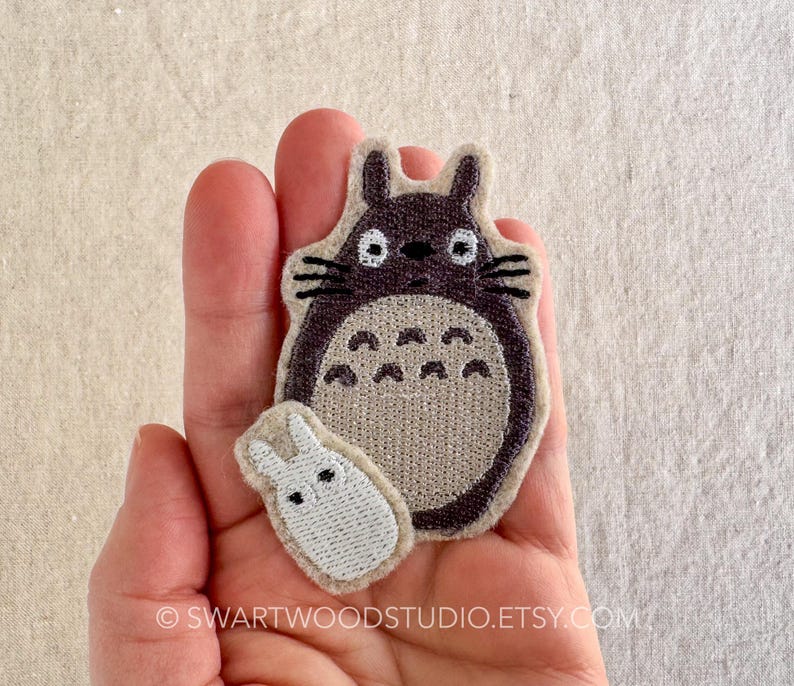 Może przedstawiać: Naszywka haftowana w kolorze brązowym i szarym przedstawiająca Totoro, postać z filmu animowanego M&oacute;j sąsiad Totoro, z mniejszą, białą haftowaną naszywką przedstawiającą kr&oacute;lika.
