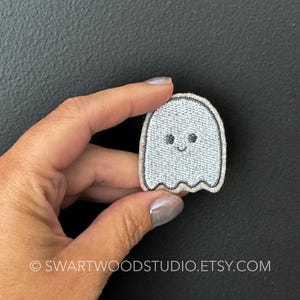 May include: A white embroidered ghost patch with a smiling face. The ghost is outlined in black thread.