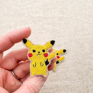 May include: A yellow embroidered iron-on patch of a cartoon Pikachu with red cheeks. The patch is held in a hand.