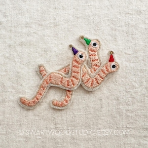 May include: Three embroidered worms wearing party hats. The worms are orange and brown and the hats are purple, green, and red.