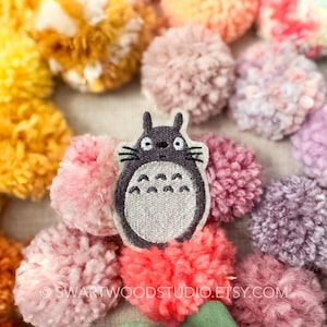 Może przedstawiać: Szara haftowana łatka z Totoro, postacią z filmu animowanego M&oacute;j sąsiad Totoro, na tle kolorowych pompon&oacute;w.