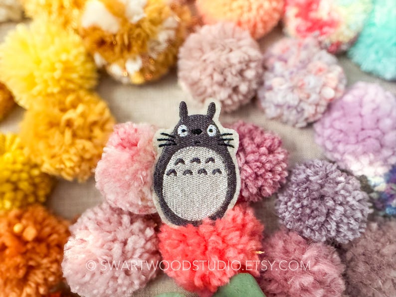 Może przedstawiać: Szara haftowana łatka z Totoro, postacią z filmu animowanego M&oacute;j sąsiad Totoro, na tle kolorowych pompon&oacute;w.