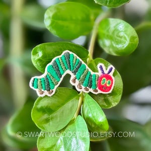 May include: A green and red embroidered iron-on patch of the very hungry caterpillar from the children's book by Eric Carle.