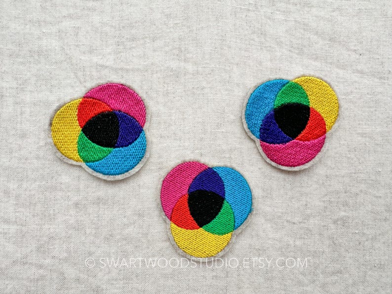 May include: Three embroidered patches in the shape of overlapping circles. The circles are in the colours of the primary colours: yellow, red, blue, and green. The centre of the patches is black.