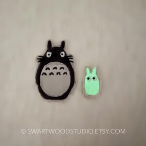 Może przedstawiać: Dwie aplikacje filcowe postaci z filmu animowanego M&oacute;j sąsiad Totoro. Większa aplikacja to czarny i szary Totoro z dużymi oczami. Mniejsza aplikacja to zielony Totoro z mniejszymi oczami.