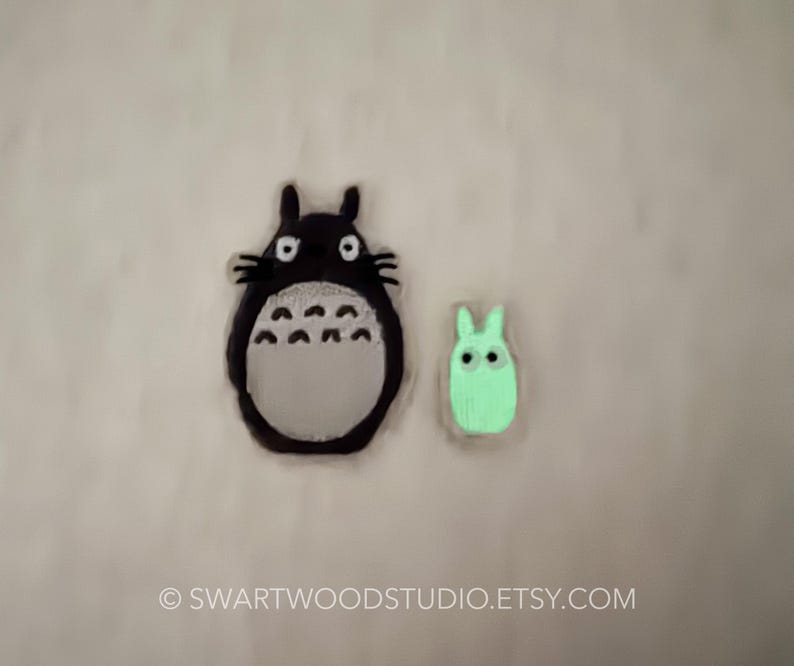 Może przedstawiać: Dwie aplikacje filcowe postaci z filmu animowanego M&oacute;j sąsiad Totoro. Większa aplikacja to czarny i szary Totoro z dużymi oczami. Mniejsza aplikacja to zielony Totoro z mniejszymi oczami.