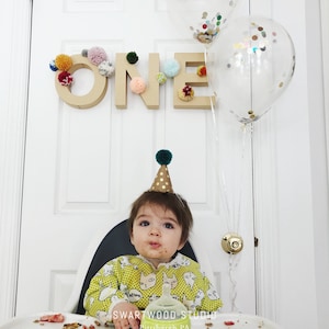 May include: A baby wearing a yellow and white shirt with a cat print and a green and gold party hat sits in a high chair. The baby has a messy face and is looking at the camera. There is a cardboard sign that says "ONE" decorated with colorful pom-poms behind the baby. There are also two clear balloons with confetti inside behind the baby.
