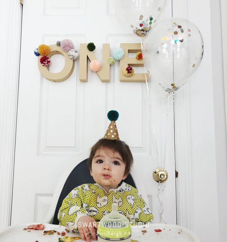 May include: A baby wearing a yellow and white shirt with a cat print and a green and gold party hat sits in a high chair. The baby has a messy face and is looking at the camera. There is a cardboard sign that says "ONE" decorated with colorful pom-poms behind the baby. There are also two clear balloons with confetti inside behind the baby.