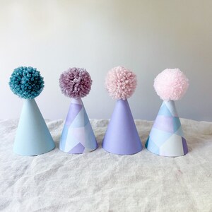May include: Four party hats with pom poms on top. The hats are blue, purple, lavender, and pink. The hats have a geometric pattern.